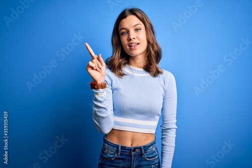 Young beautiful blonde girl wearing casual sweater standing over blue isolated background showing and pointing up with finger number one while smiling confident and happy.