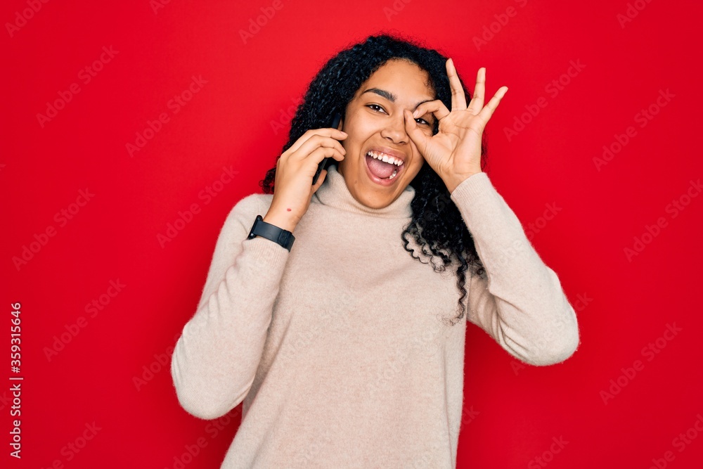 Young african american curly woman having conversation talking on the smartphone with happy face smiling doing ok sign with hand on eye looking through fingers Young african american curly woman having conversation talking on the smartphone with happy face smiling doing ok sign with hand on eye looking through fingers