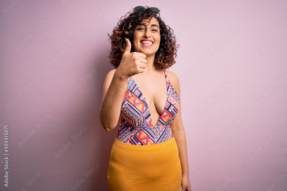 Young beautiful arab woman on vacation wearing swimsuit and sunglasses over pink background doing happy thumbs up gesture with hand. Approving expression looking at the camera showing success. Young beautiful arab woman on vacation wearing swimsuit and sunglasses over pink background doing happy thumbs up gesture with hand. Approving expression looking at the camera showing success.