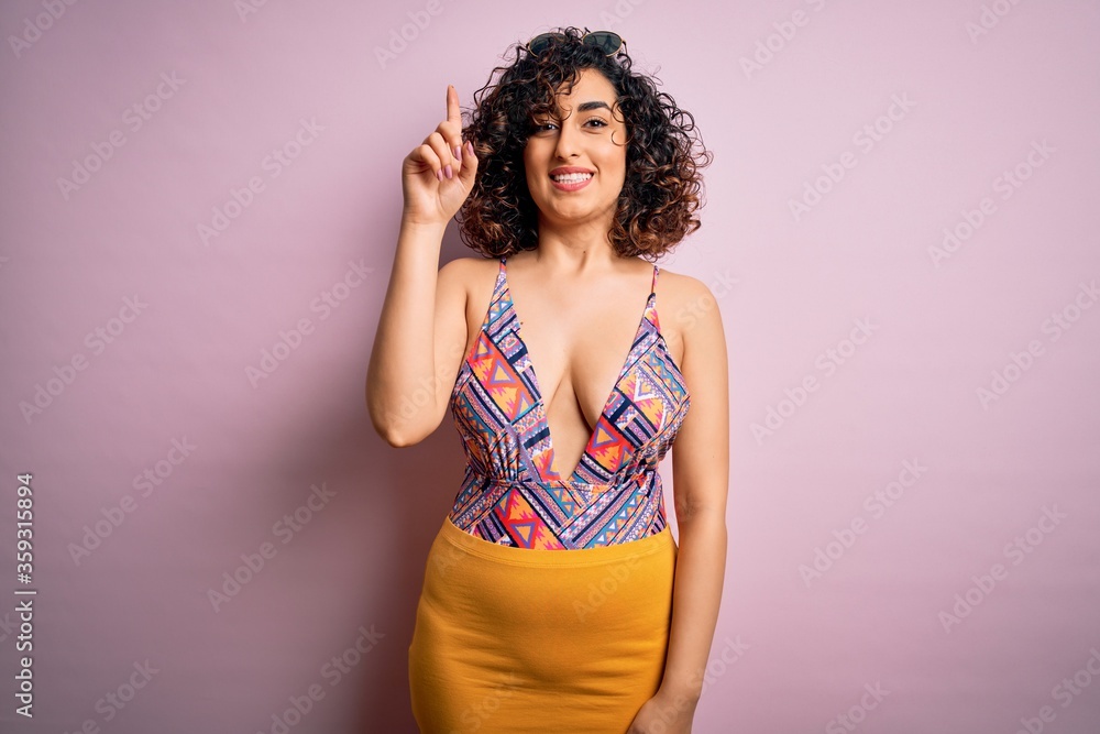 Young beautiful arab woman on vacation wearing swimsuit and sunglasses over pink background showing and pointing up with finger number one while smiling confident and happy. Young beautiful arab woman on vacation wearing swimsuit and sunglasses over pink background showing and pointing up with finger number one while smiling confident and happy.