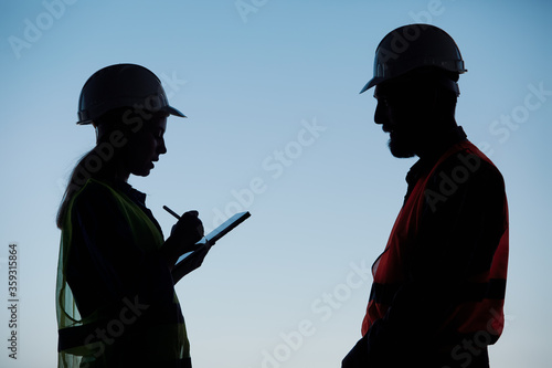 Silhouettes of an engineer with a tablet in his hands and a construction worker in a hard hat against the blue sky stand opposite each other