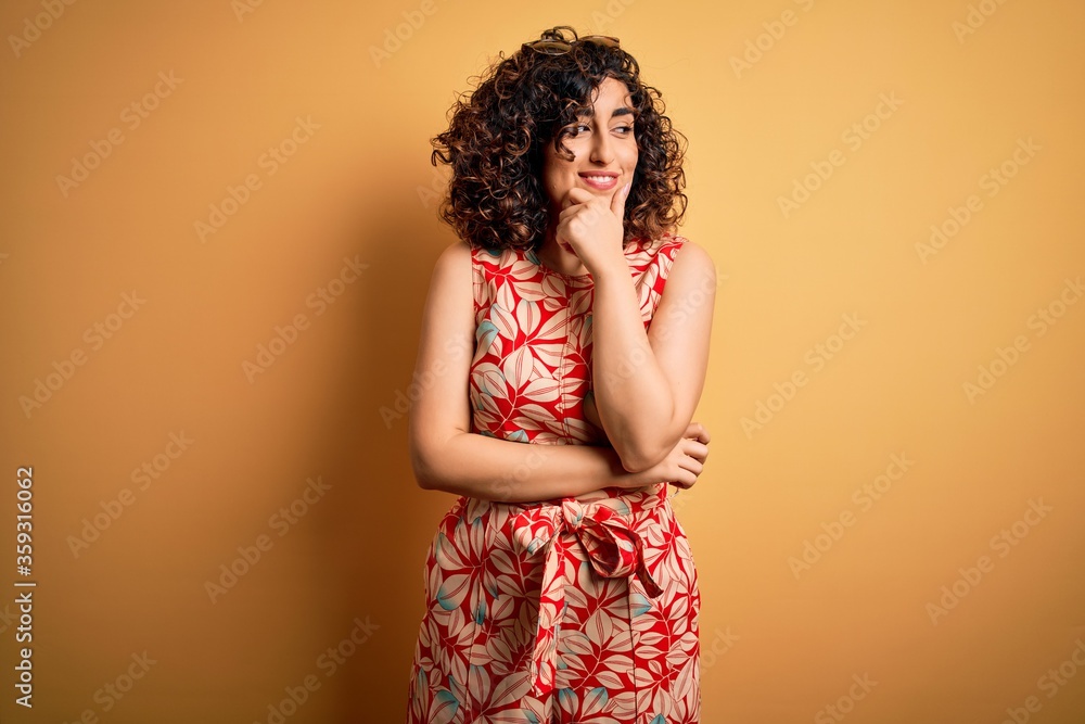 Young beautiful curly arab woman on vacation wearing summer floral dress and sunglasses with hand on chin thinking about question, pensive expression. Smiling with thoughtful face. Doubt concept. Young beautiful curly arab woman on vacation wearing summer floral dress and sunglasses with hand on chin thinking about question, pensive expression. Smiling with thoughtful face. Doubt concept.
