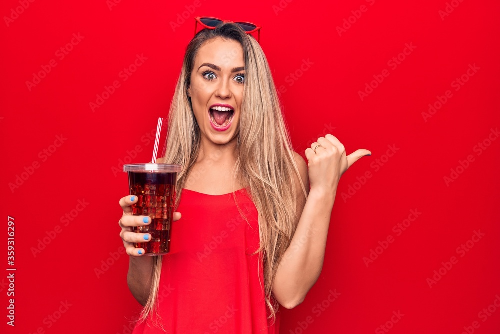 Beautiful blonde woman drinking cola beverage refreshment using straw over red background pointing thumb up to the side smiling happy with open mouth - obrazy, fototapety, plakaty 