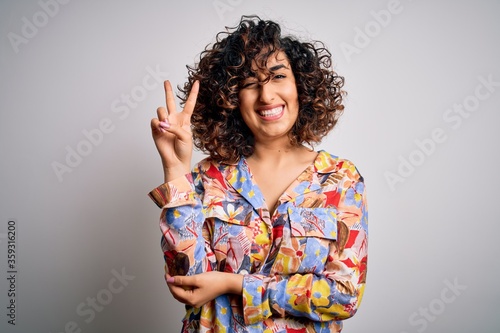 Young beautiful curly arab woman wearing floral colorful shirt standing over white background smiling with happy face winking at the camera doing victory sign. Number two.