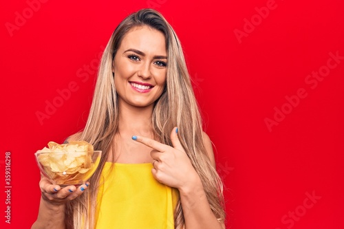 Young beautiful blonde woman holding bowl with potato chips over isolated red background smiling happy pointing with hand and finger