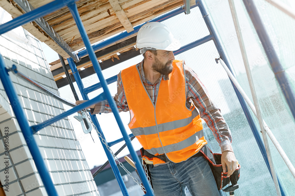 A stylish Builder with a beard in an orange vest with a safety belt conducts construction work