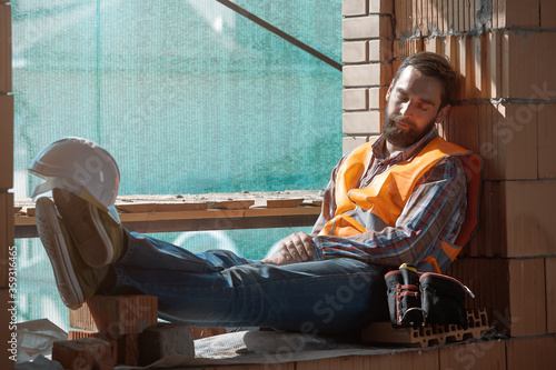 A stylish Builder with a beard in an orange vest with a safety belt sleeps in the workplace