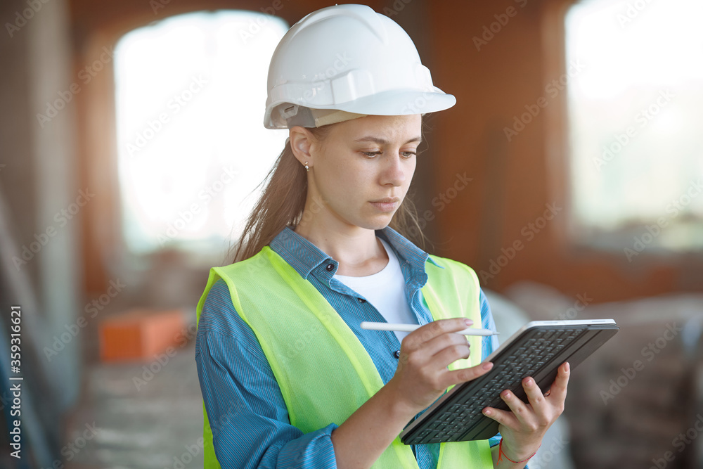 Fototapeta premium Absorbed in the work of a woman engineer working with a tablet on the background of the construction site. Portrait of a young architect, protective equipment. Selective focus.