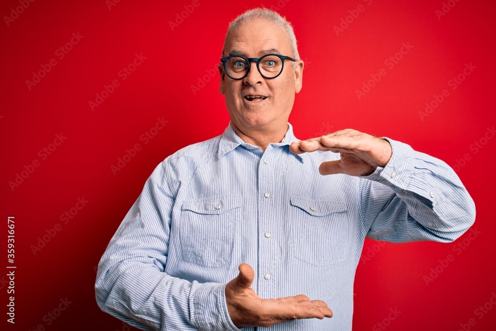 Middle age handsome hoary man wearing casual striped shirt and glasses over red background gesturing with hands showing big and large size sign, measure symbol. Smiling looking at the camera. Measure Middle age handsome hoary man wearing casual striped shirt and glasses over red background gesturing with hands showing big and large size sign, measure symbol. Smiling looking at the camera. Measure