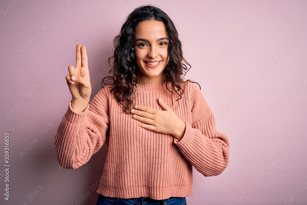 Young beautiful woman with curly hair wearing casual sweater over isolated pink background smiling swearing with hand on chest and fingers up, making a loyalty promise oath Young beautiful woman with curly hair wearing casual sweater over isolated pink background smiling swearing with hand on chest and fingers up, making a loyalty promise oath