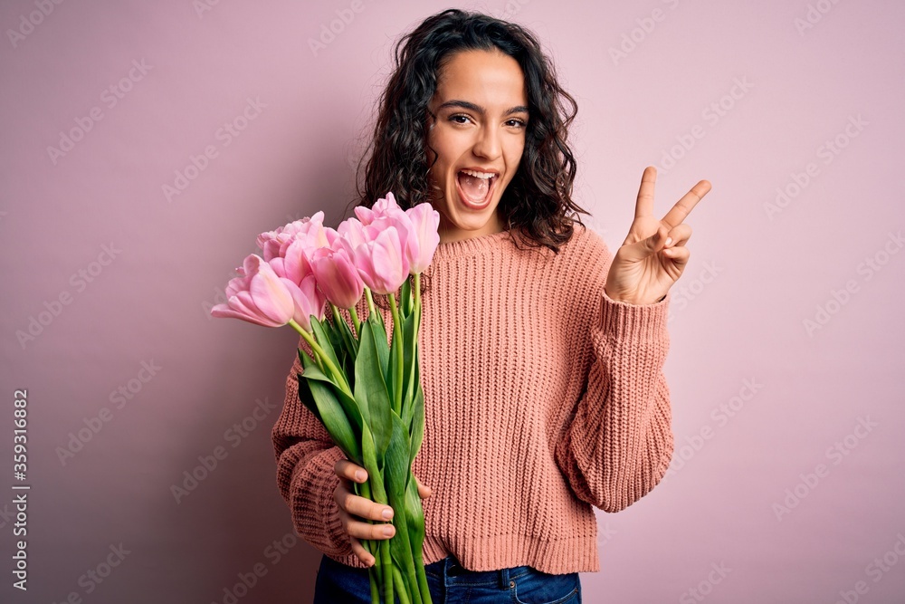Young beautiful romantic woman with curly hair holding bouquet of pink tulips smiling with happy face winking at the camera doing victory sign with fingers. Number two. Young beautiful romantic woman with curly hair holding bouquet of pink tulips smiling with happy face winking at the camera doing victory sign with fingers. Number two.