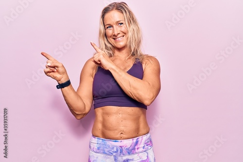 Middle age caucasian blonde woman wearing sportswear over pink background smiling and looking at the camera pointing with two hands and fingers to the side.