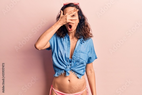 Young beautiful hispanic woman wearing bikini and shirt peeking in shock covering face and eyes with hand, looking through fingers afraid