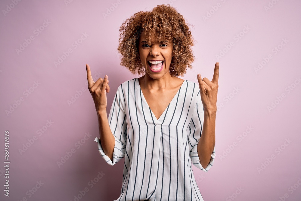 Beautiful african american woman with curly hair wearing striped t-shirt over pink background shouting with crazy expression doing rock symbol with hands up. Music star. Heavy concept. Beautiful african american woman with curly hair wearing striped t-shirt over pink background shouting with crazy expression doing rock symbol with hands up. Music star. Heavy concept.