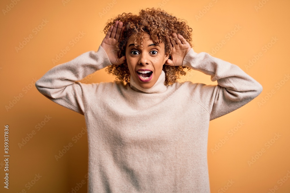 Young beautiful african american woman wearing turtleneck sweater over yellow background Smiling cheerful playing peek a boo with hands showing face. Surprised and exited Young beautiful african american woman wearing turtleneck sweater over yellow background Smiling cheerful playing peek a boo with hands showing face. Surprised and exited