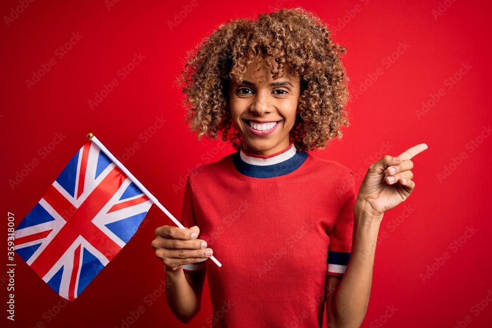 Young african american curly woman holding uk flag celebrating brexit referendum very happy pointing with hand and finger to the side Young african american curly woman holding uk flag celebrating brexit referendum very happy pointing with hand and finger to the side