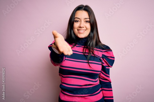 Young brunette elegant woman wearing striped shirt over pink isolated background smiling friendly offering handshake as greeting and welcoming. Successful business.