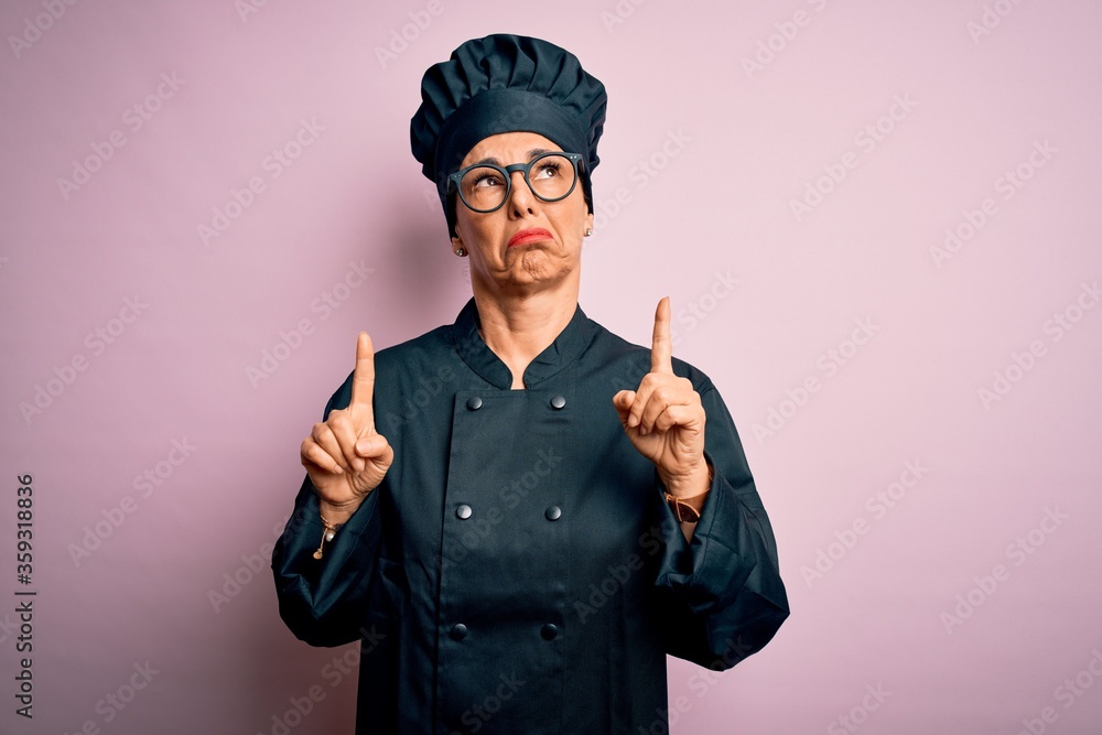 Middle age brunette chef woman wearing cooker uniform and hat over isolated pink background Pointing up looking sad and upset, indicating direction with fingers, unhappy and depressed. Middle age brunette chef woman wearing cooker uniform and hat over isolated pink background Pointing up looking sad and upset, indicating direction with fingers, unhappy and depressed.