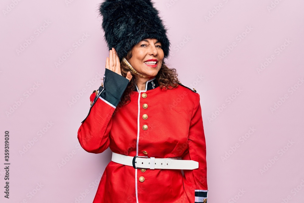 Obraz premium Middle age beautiful wales guard woman wearing traditional uniform over pink background smiling with hand over ear listening an hearing to rumor or gossip. Deafness concept.