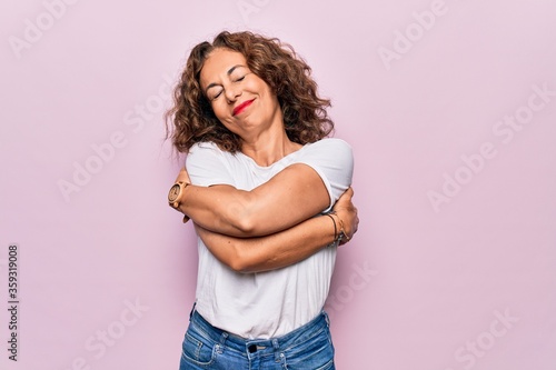 Tela Middle age beautiful woman wearing casual t-shirt standing over isolated pink background hugging oneself happy and positive, smiling confident