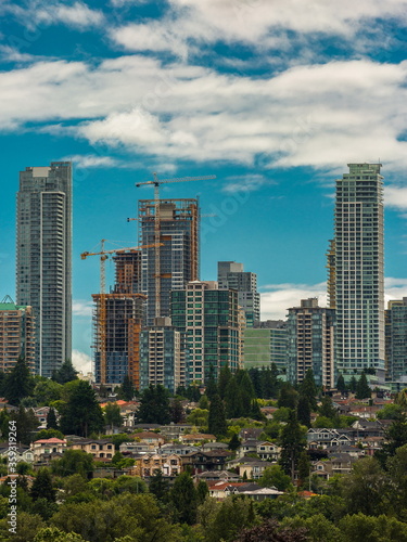 Construction of New Residential District in the city of Burnaby, high-rise buildings under construction and construction cranes against the backdrop of blue cloudy sky and village in the foreground