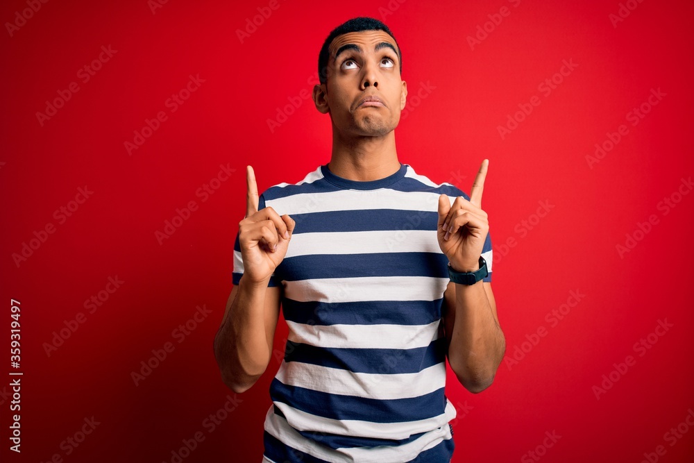 Handsome african american man wearing casual striped t-shirt standing over red background Pointing up looking sad and upset, indicating direction with fingers, unhappy and depressed. Handsome african american man wearing casual striped t-shirt standing over red background Pointing up looking sad and upset, indicating direction with fingers, unhappy and depressed.