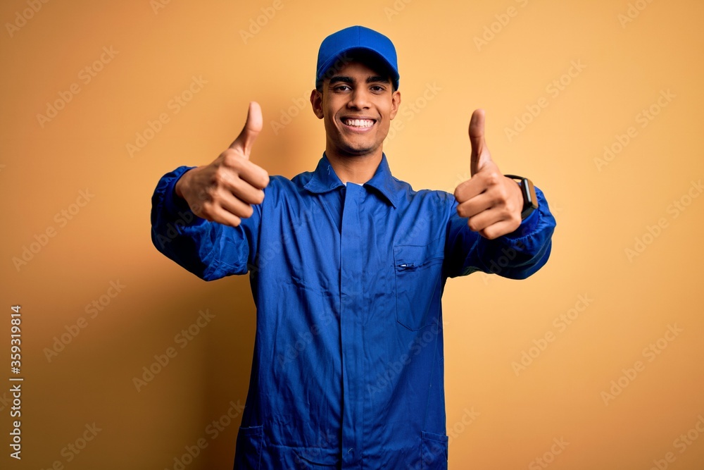 Young african american mechanic man wearing blue uniform and cap over yellow background approving doing positive gesture with hand, thumbs up smiling and happy for success. Winner gesture.