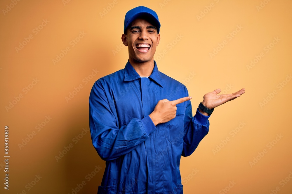 Young african american mechanic man wearing blue uniform and cap over yellow background amazed and smiling to the camera while presenting with hand and pointing with finger.