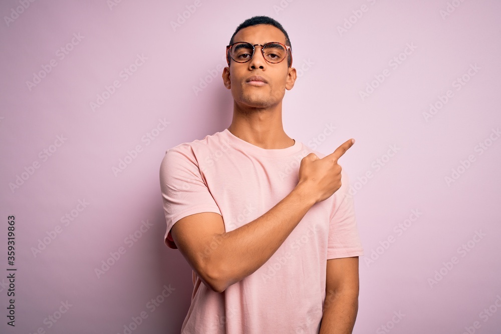 Handsome african american man wearing casual t-shirt and glasses over pink background Pointing with hand finger to the side showing advertisement, serious and calm face Handsome african american man wearing casual t-shirt and glasses over pink background Pointing with hand finger to the side showing advertisement, serious and calm face
