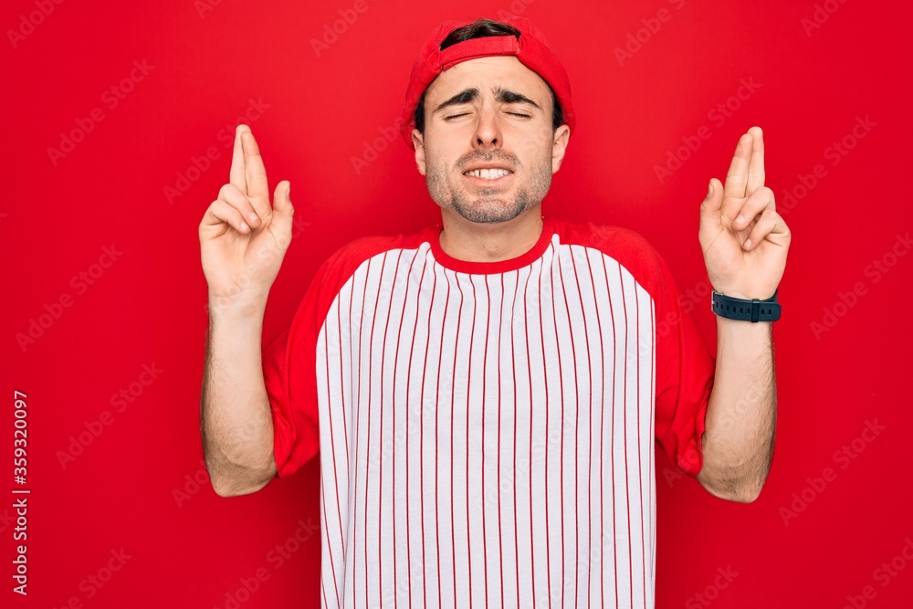 Young handsome sporty man with blue eyes wearing striped baseball t-shirt and cap gesturing finger crossed smiling with hope and eyes closed. Luck and superstitious concept. Young handsome sporty man with blue eyes wearing striped baseball t-shirt and cap gesturing finger crossed smiling with hope and eyes closed. Luck and superstitious concept.