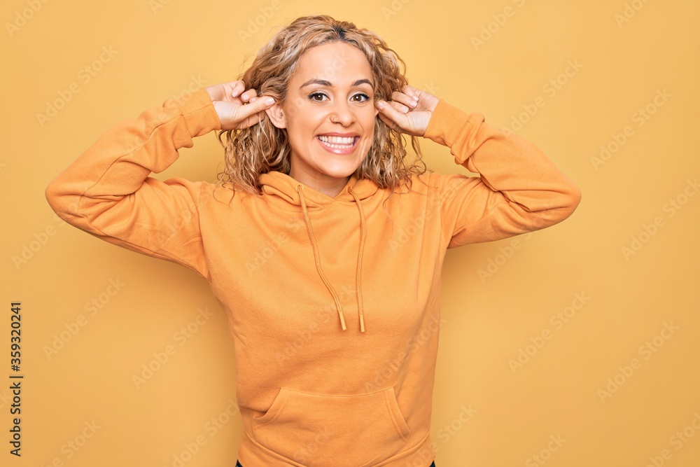 Young beautiful blonde sporty woman wearing casual sweatshirt over yellow background Smiling pulling ears with fingers, funny gesture. Audition problem Young beautiful blonde sporty woman wearing casual sweatshirt over yellow background Smiling pulling ears with fingers, funny gesture. Audition problem