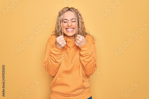 Young beautiful blonde sporty woman wearing casual sweatshirt over yellow background excited for success with arms raised and eyes closed celebrating victory smiling. Winner concept.