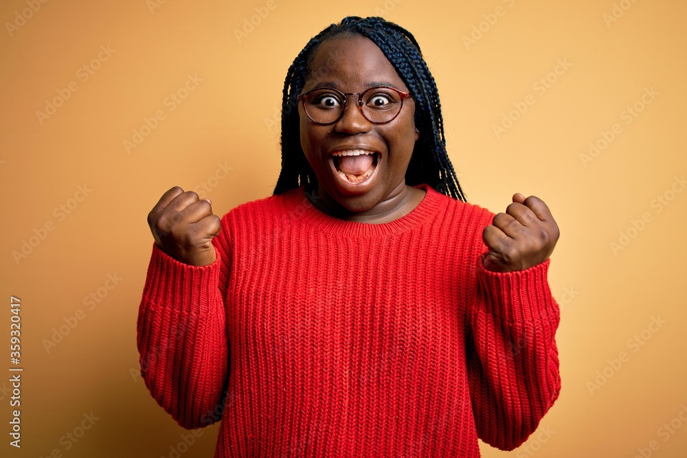 African american plus size woman with braids wearing casual sweater over yellow background celebrating surprised and amazed for success with arms raised and open eyes. Winner concept. African american plus size woman with braids wearing casual sweater over yellow background celebrating surprised and amazed for success with arms raised and open eyes. Winner concept.