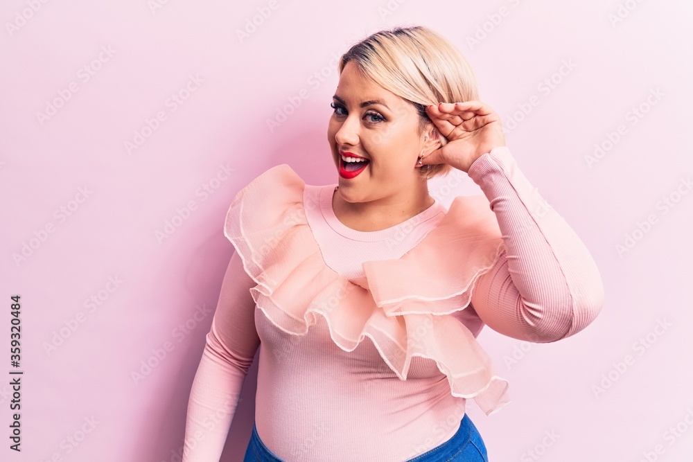 Young beautiful blonde plus size woman wearing casual sweater over isolated pink background smiling with hand over ear listening and hearing to rumor or gossip. Deafness concept. Young beautiful blonde plus size woman wearing casual sweater over isolated pink background smiling with hand over ear listening and hearing to rumor or gossip. Deafness concept.