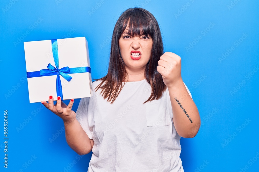 Young plus size woman holding gift annoyed and frustrated shouting with anger, yelling crazy with anger and hand raised Young plus size woman holding gift annoyed and frustrated shouting with anger, yelling crazy with anger and hand raised