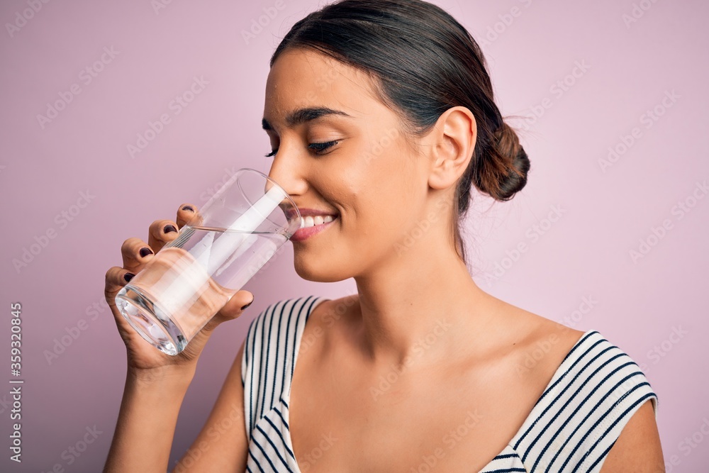 Young beautiful brunette woman smiling happy and confident. Standing with smile on face drinking glass of healthy water to refreshment over isolated pink background Young beautiful brunette woman smiling happy and confident. Standing with smile on face drinking glass of healthy water to refreshment over isolated pink background