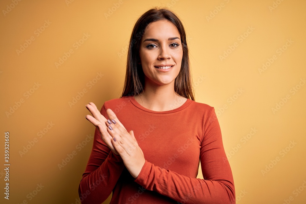 Obraz premium Young beautiful brunette woman wearing casual t-shirt standing over yellow background clapping and applauding happy and joyful, smiling proud hands together