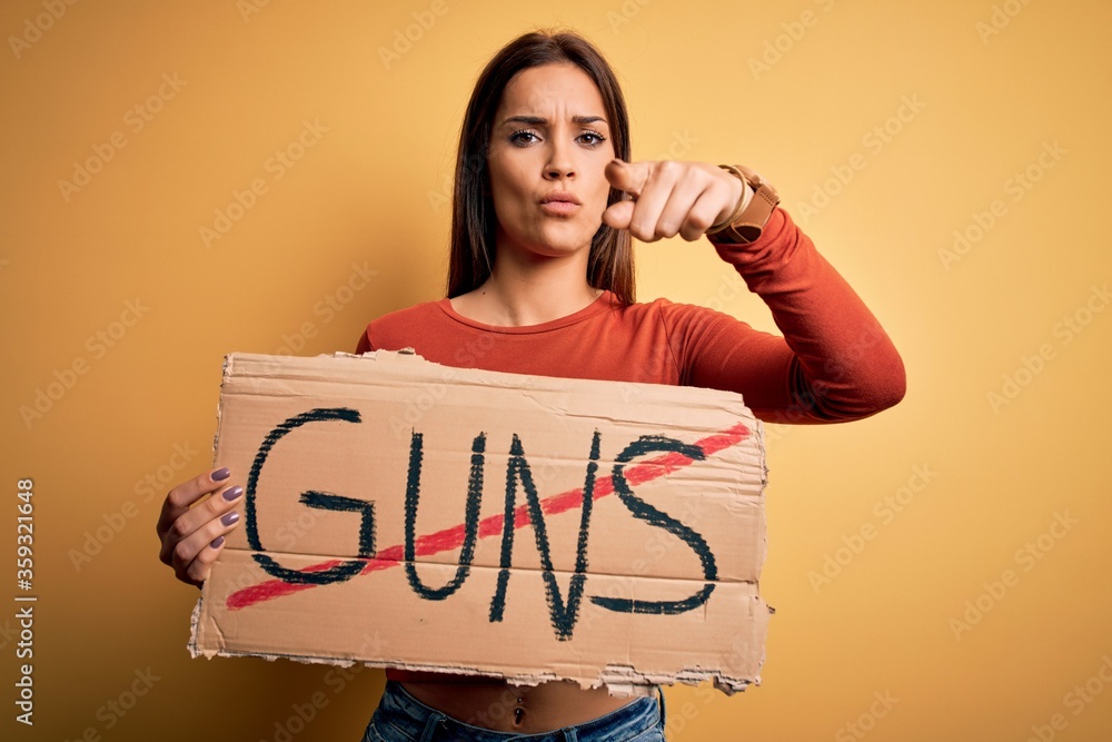 Young beautiful activist woman asking for peace holding banner with stop guns message pointing with finger to the camera and to you, hand sign, positive and confident gesture from the front Young beautiful activist woman asking for peace holding banner with stop guns message pointing with finger to the camera and to you, hand sign, positive and confident gesture from the front