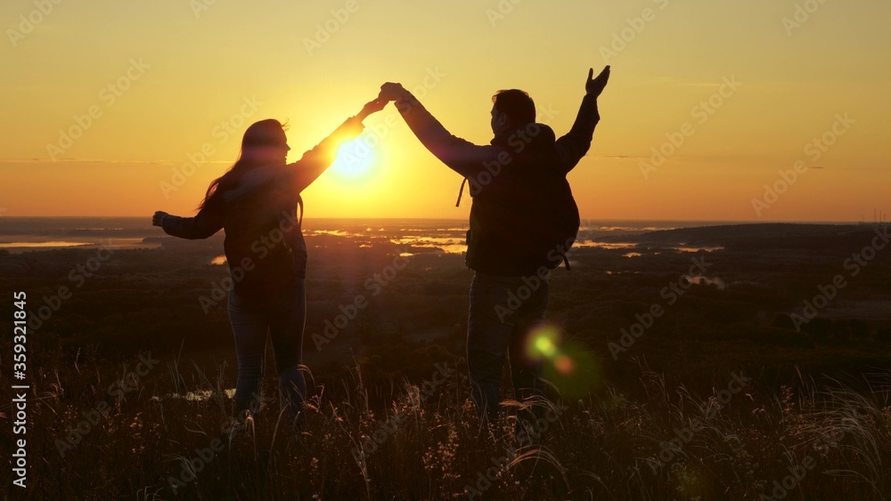 Travelers, man and woman with backpack, go to edge of hill in rays of dawn, raise their hands, clap their hands and enjoy victory and beautiful sun and landscape. family travel and tourism concept