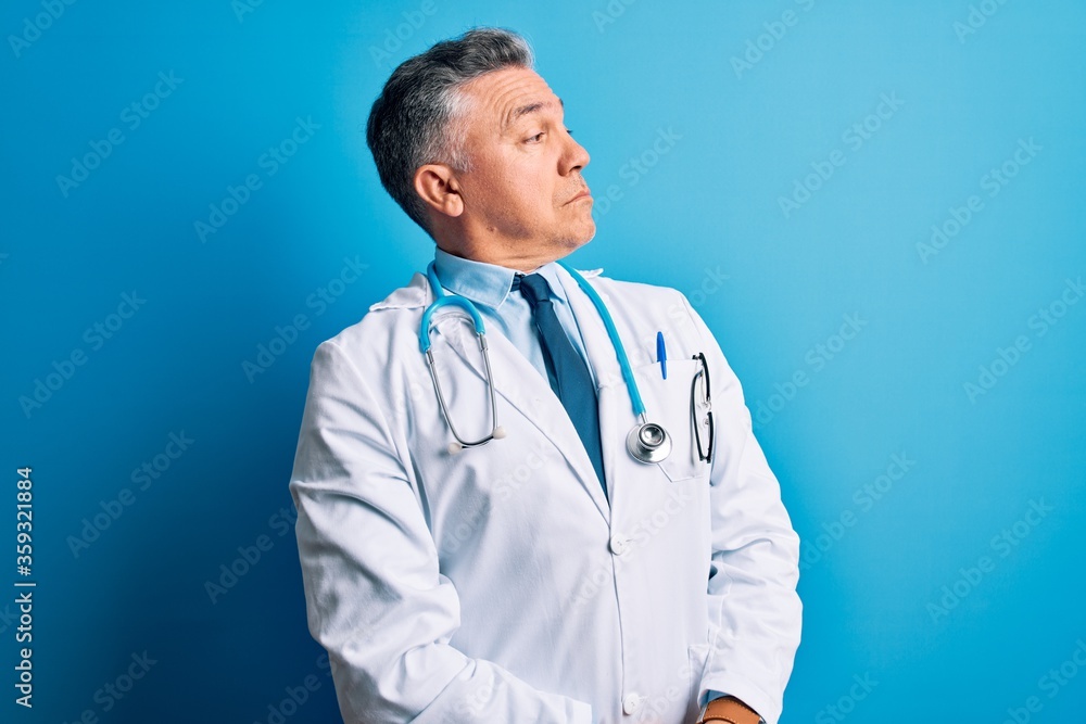 Middle age handsome grey-haired doctor man wearing coat and blue stethoscope looking to the side with arms crossed convinced and confident Middle age handsome grey-haired doctor man wearing coat and blue stethoscope looking to the side with arms crossed convinced and confident