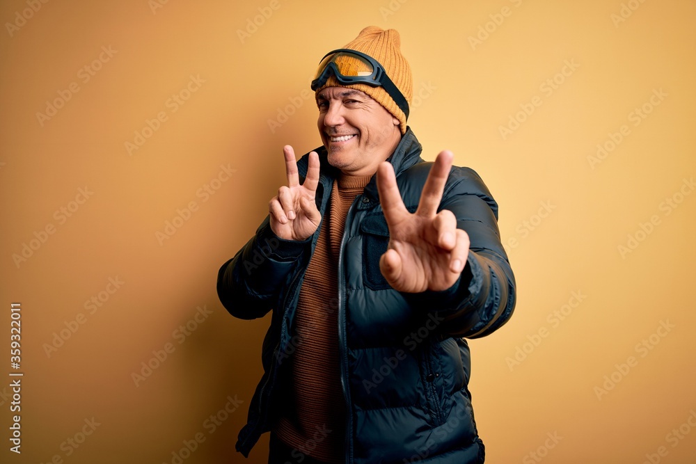 Middle age handsome grey-haired skier man on vacation wearing ski goggles smiling looking to the camera showing fingers doing victory sign. Number two. Middle age handsome grey-haired skier man on vacation wearing ski goggles smiling looking to the camera showing fingers doing victory sign. Number two.