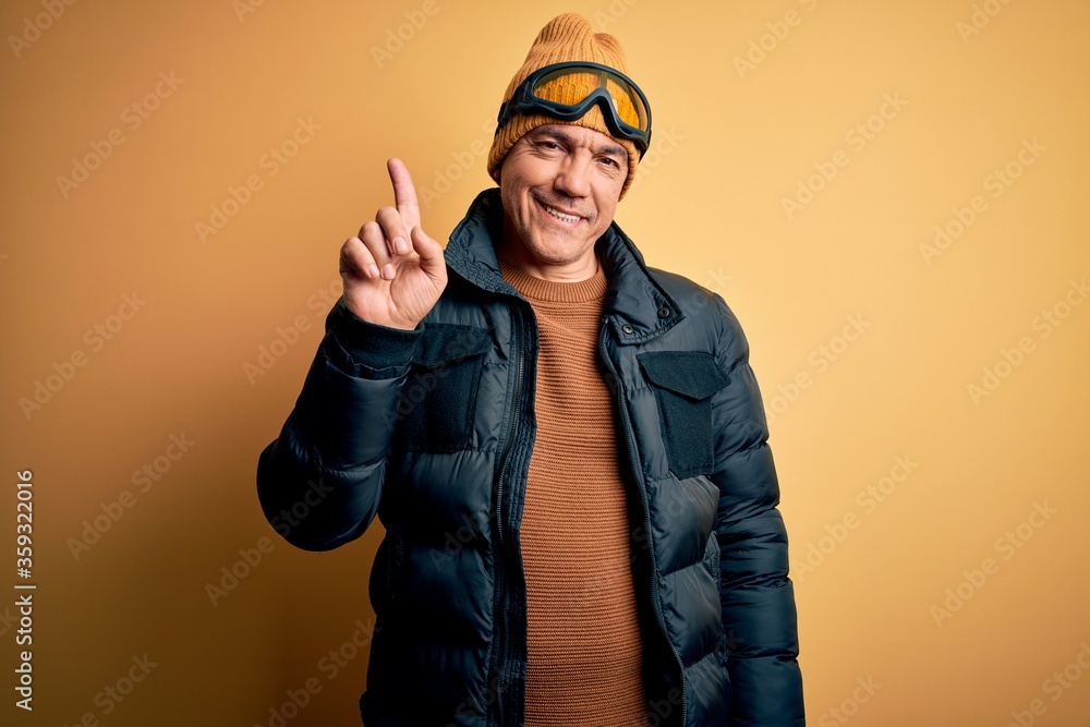 Middle age handsome grey-haired skier man on vacation wearing ski goggles showing and pointing up with finger number one while smiling confident and happy. Middle age handsome grey-haired skier man on vacation wearing ski goggles showing and pointing up with finger number one while smiling confident and happy.
