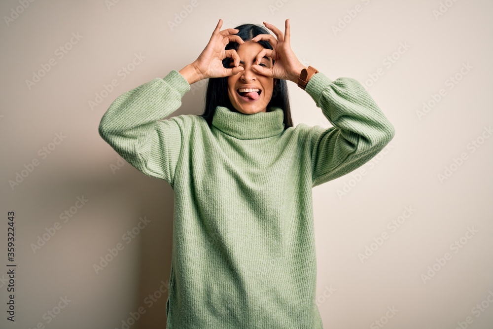 Young beautiful hispanic woman wearing green winter sweater over isolated background doing ok gesture like binoculars sticking tongue out, eyes looking through fingers. Crazy expression. Young beautiful hispanic woman wearing green winter sweater over isolated background doing ok gesture like binoculars sticking tongue out, eyes looking through fingers. Crazy expression.