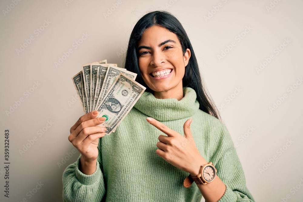 Young beautiful hispanic woman holding a pack of one dollar bunch of dollars very happy pointing with hand and finger