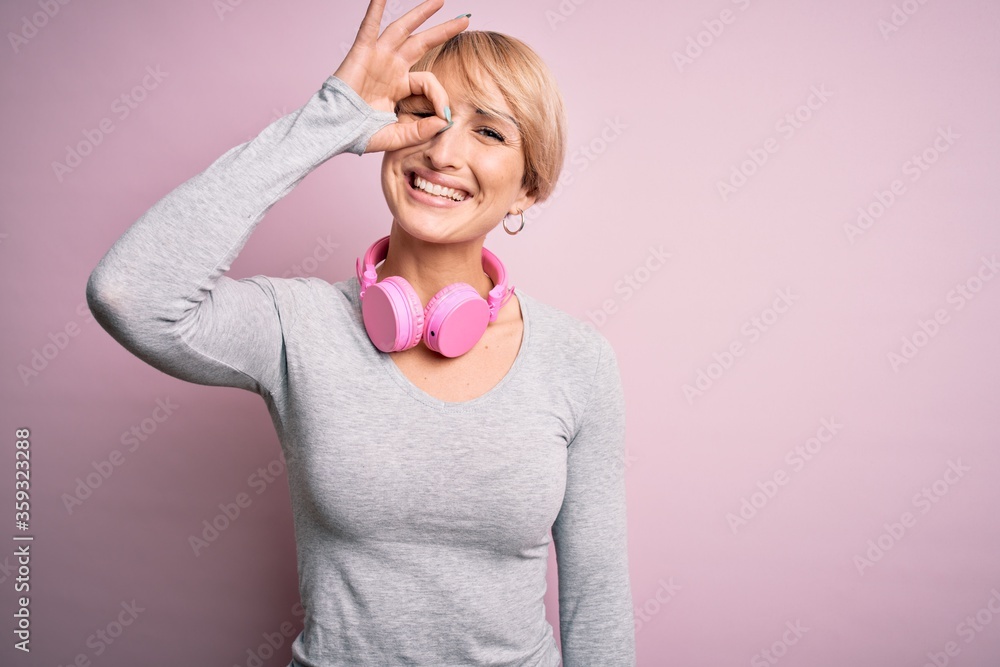 Young blonde woman with short hair wearing headphones on neck over pink background doing ok gesture with hand smiling, eye looking through fingers with happy face. Young blonde woman with short hair wearing headphones on neck over pink background doing ok gesture with hand smiling, eye looking through fingers with happy face.
