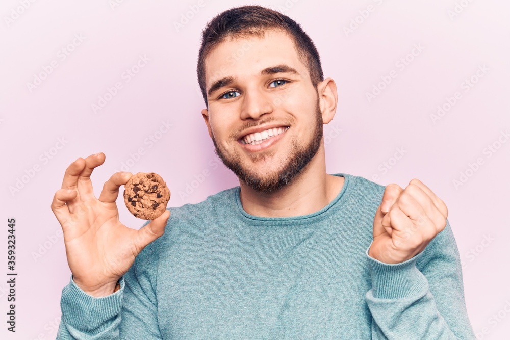 Young handsome man holding cookie screaming proud, celebrating victory and success very excited with raised arm Young handsome man holding cookie screaming proud, celebrating victory and success very excited with raised arm