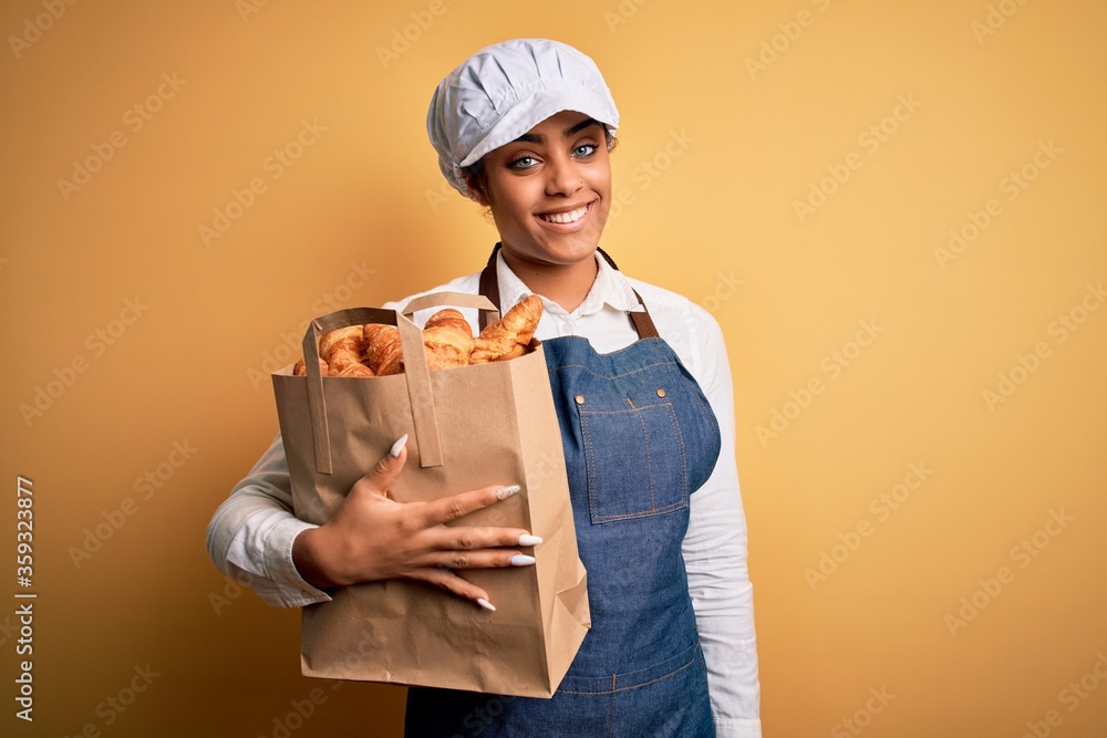 Young african american baker girl wearing apron holding bag with french croissants with a happy face standing and smiling with a confident smile showing teeth Young african american baker girl wearing apron holding bag with french croissants with a happy face standing and smiling with a confident smile showing teeth