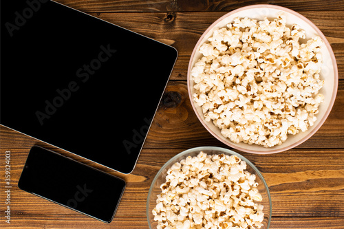 Bowls filled with modern tablet computer, phone and popcorn eaten while watching movies and TV series on wooden floor.  Phone and Tablet mockup with black copy space.