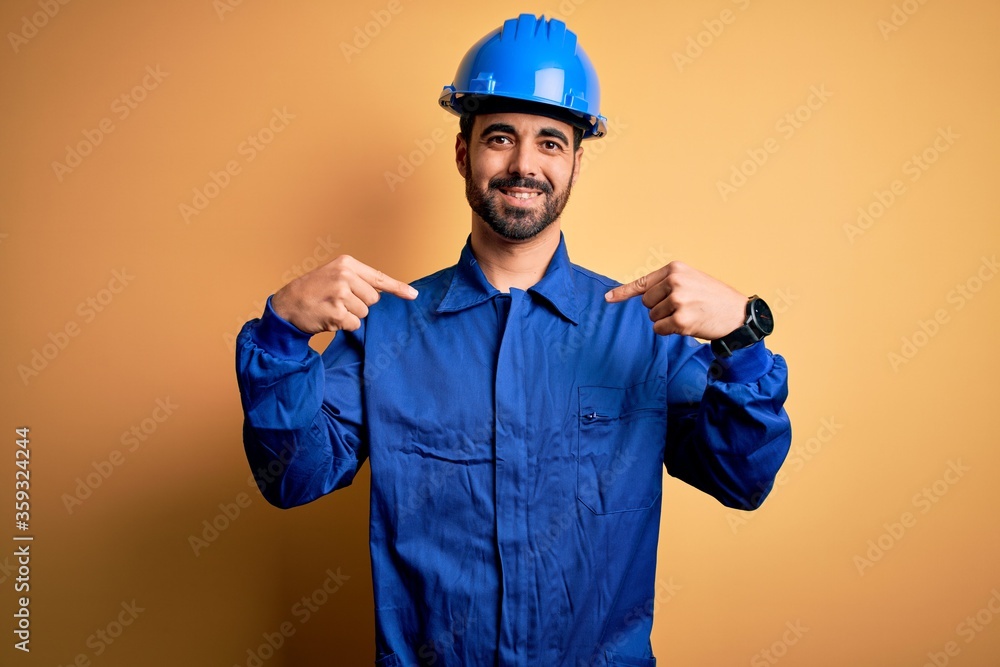 Mechanic man with beard wearing blue uniform and safety helmet over yellow background looking confident with smile on face, pointing oneself with fingers proud and happy.