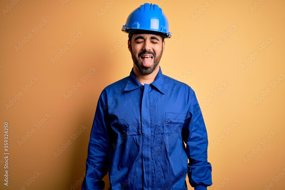 Mechanic man with beard wearing blue uniform and safety helmet over yellow background sticking tongue out happy with funny expression. Emotion concept.